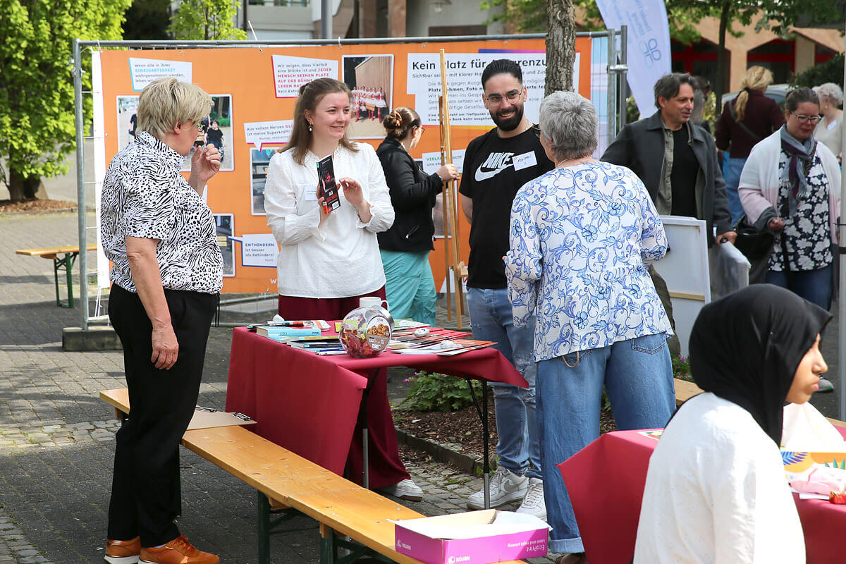 An einem Informationsstand im Freien, an dem Broschüren und Plakate ausliegen, stehen Menschen und unterhalten sich. Andere unterhalten sich in der Nähe. Die Szene scheint eine Gemeinschaftsveranstaltung oder ein Fest zu sein.