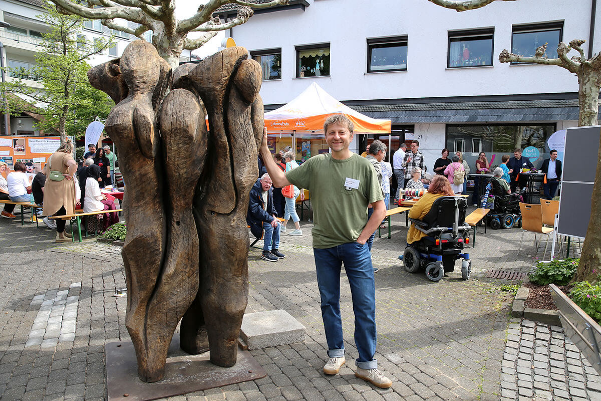 Eine Person in einem grünen Hemd steht neben einer großen Holzskulptur auf einem Platz im Freien, im Hintergrund sitzen Menschen an Tischen und unter Zelten.