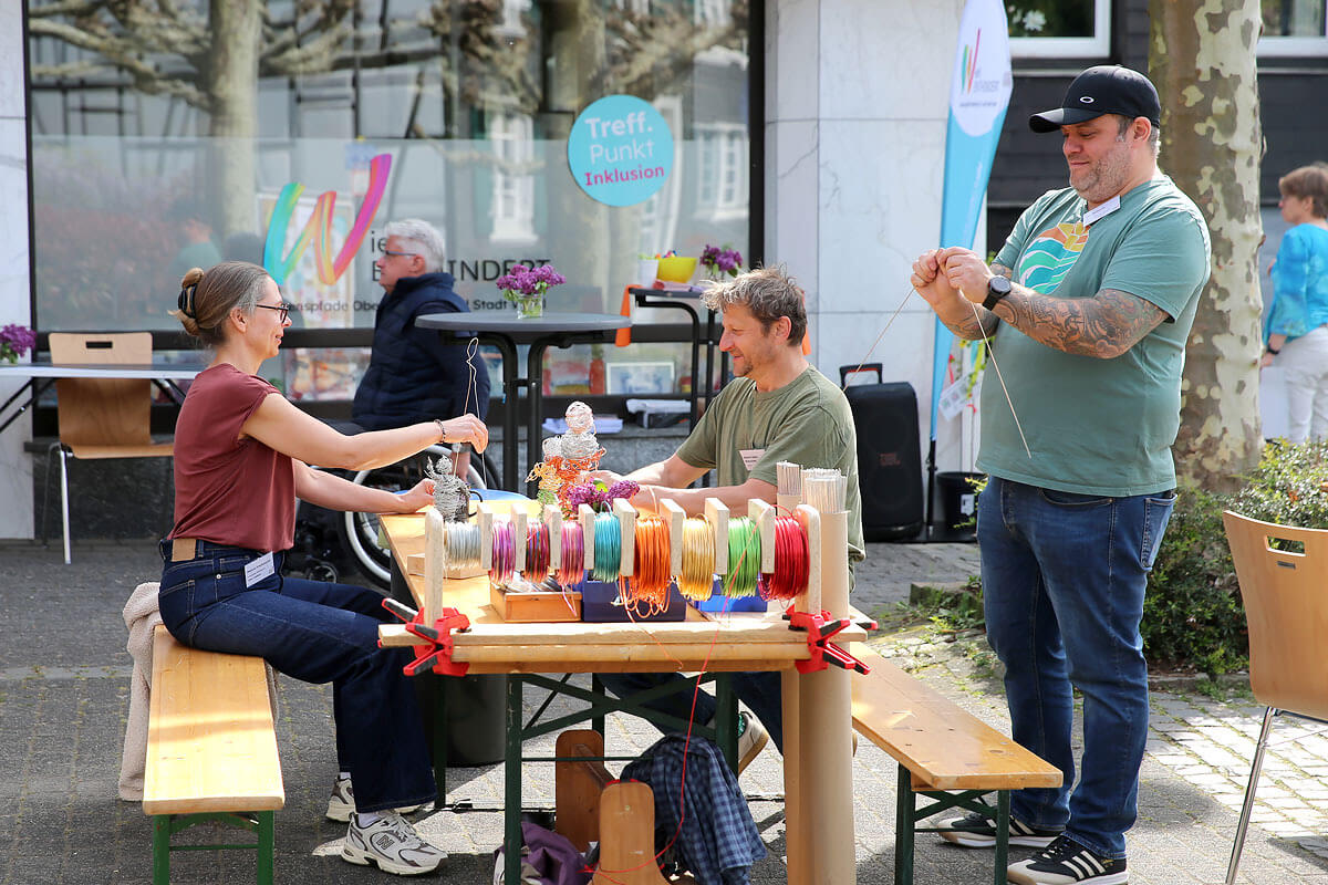 Drei Erwachsene arbeiten an einem Tisch im Freien mit bunten Fäden; zwei sitzen und einer steht, im Hintergrund sind ein Gebäude und Bäume zu sehen.