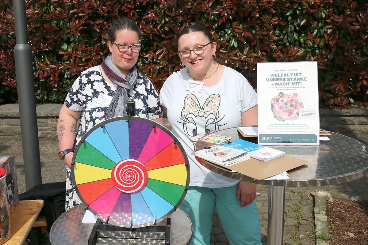 Zwei Frauen stehen an einem Tisch im Freien, auf dem ein buntes Glücksrad, Informationsbroschüren und ein Schild zur Förderung der Vielfalt zu sehen sind.