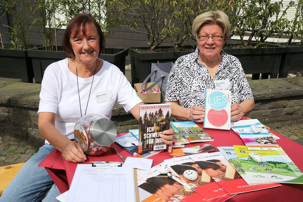 Zwei Frauen sitzen an einem Tisch im Freien, auf dem Bücher, Broschüren, eine Anmeldeliste, Werbegeschenke und Informationsmaterial ausliegen.