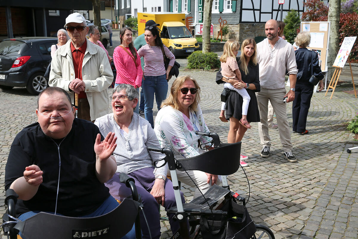Eine Gruppe von Menschen, darunter auch Personen mit Mobilitätshilfen, versammelt sich im Freien auf einer Kopfsteinpflasterstraße in einer Kleinstadtkulisse.