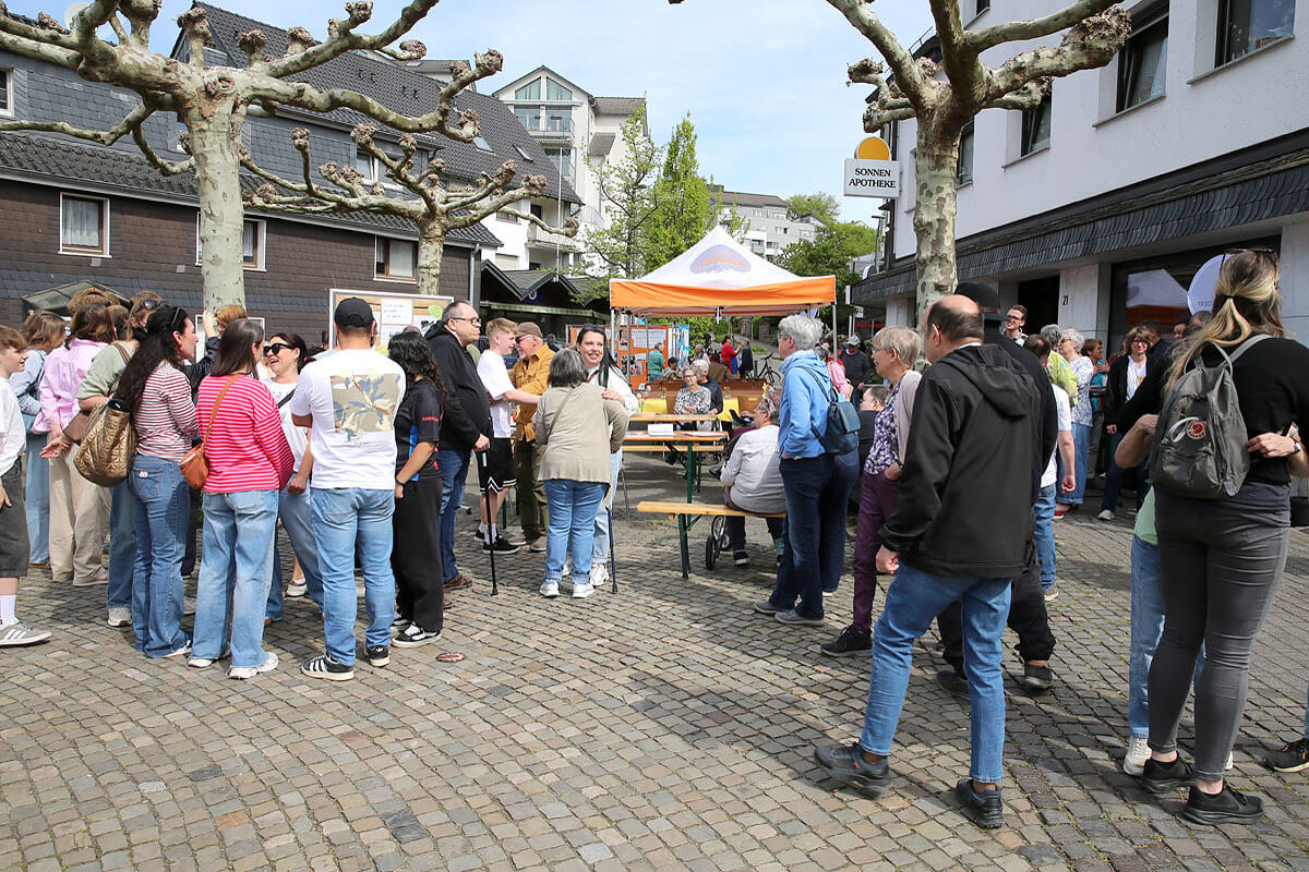 Eine Gruppe von Menschen steht unter einem orangefarbenen Vordach auf einem gepflasterten Außenbereich mit Bäumen und Gebäuden im Hintergrund und unterhält sich an einem Tisch, auf dem Essen steht.