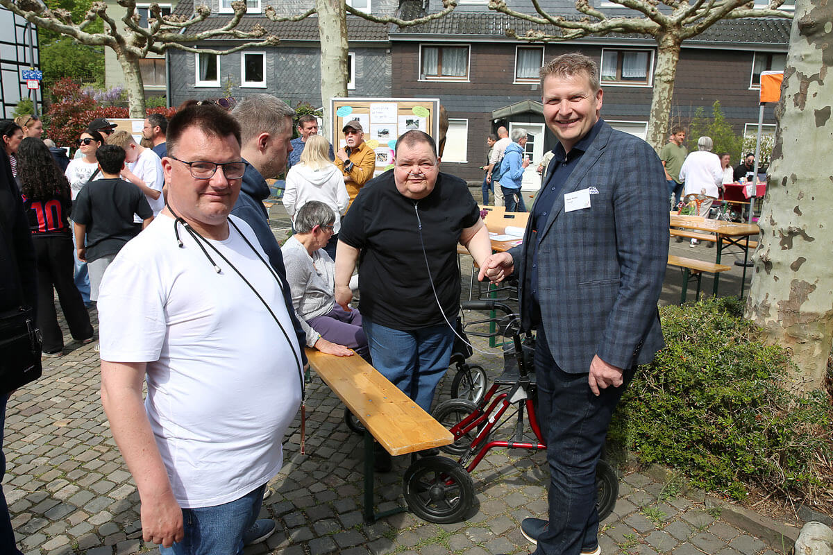 Drei Männer, von denen einer eine Gehhilfe benutzt, posieren gemeinsam bei einer Gemeinschaftsveranstaltung im Freien, im Hintergrund sind Menschen und Informationsstände zu sehen.