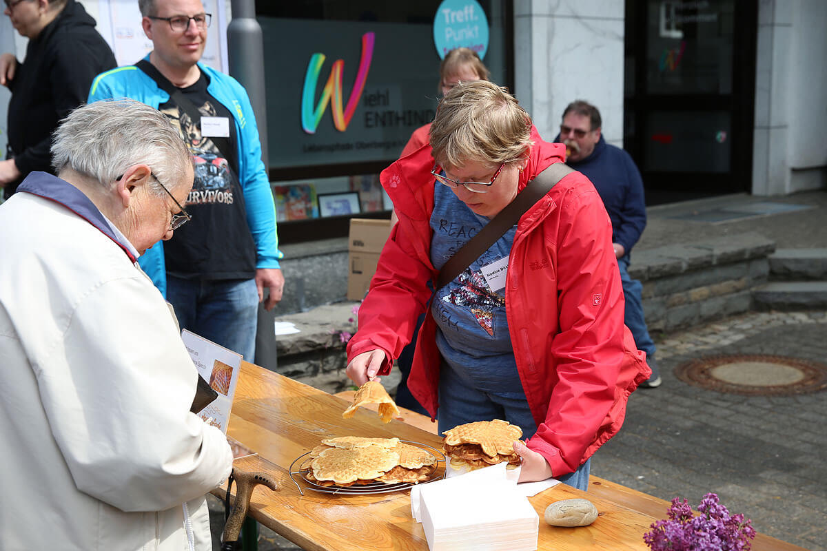 Eine Frau in einer roten Jacke serviert an einem Tisch im Freien Waffeln, während andere in der Nähe stehen. Auf dem Tisch liegen Zeitungen und eine Schachtel.