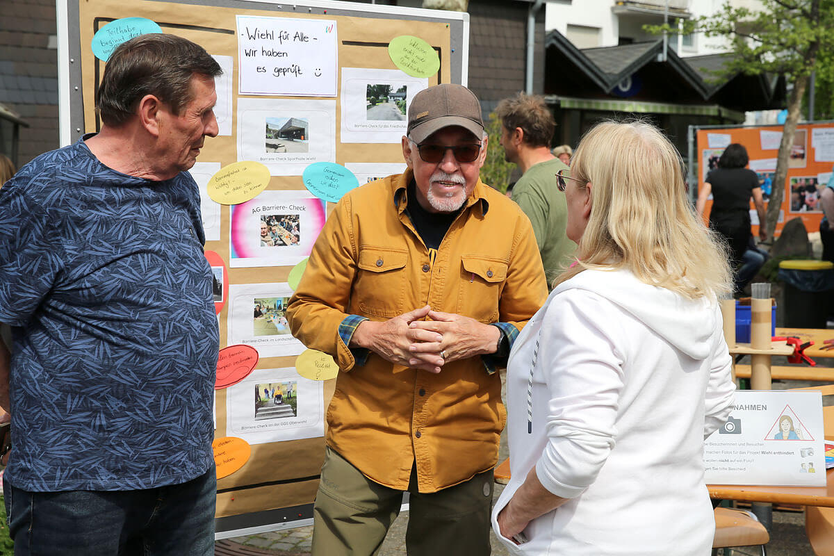 Bei einer Veranstaltung im Freien stehen drei Erwachsene vor Anschlagtafeln, an denen Zettel, Fotos und schriftliche Informationen aushängen, und unterhalten sich.