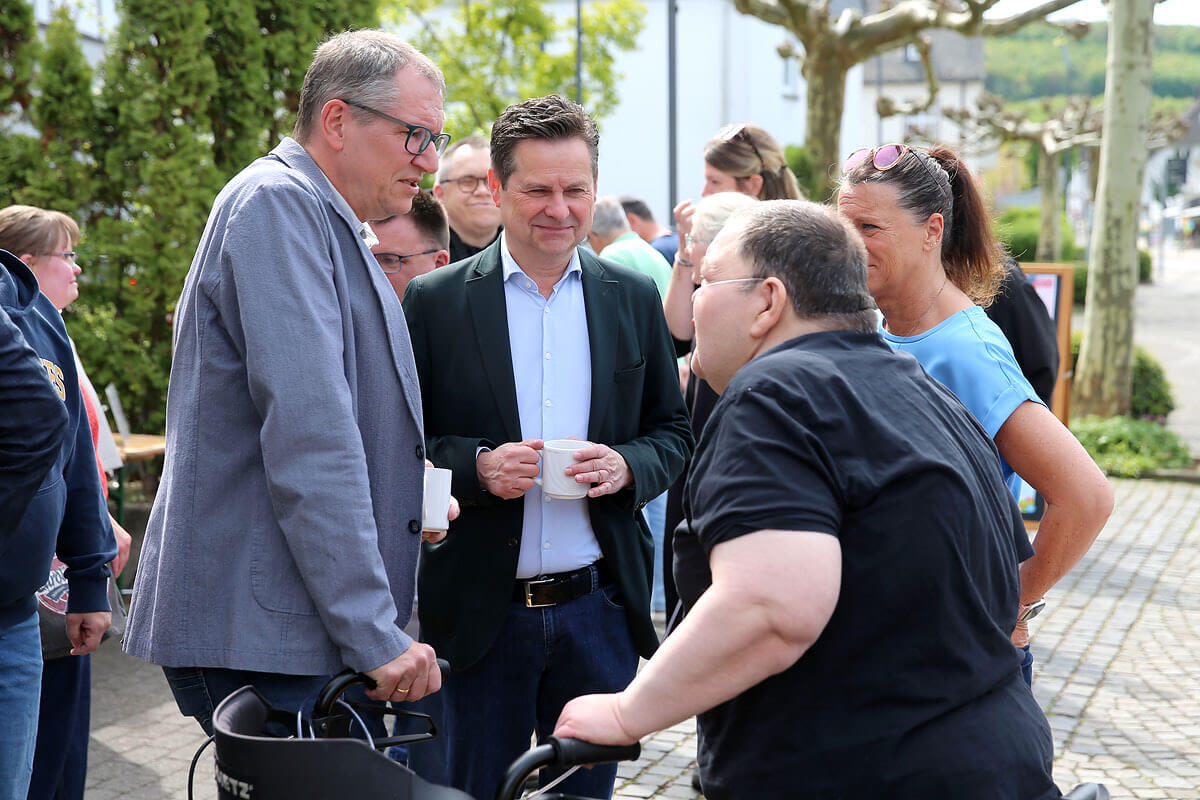 Eine Gruppe von Menschen steht im Freien und unterhält sich. Ein Mann hält einen Becher in der Hand, während ein anderer eine Gehhilfe benutzt. Im Hintergrund sind Bäume und gepflasterter Boden zu sehen.