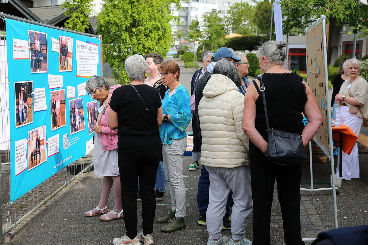 Eine Gruppe von Menschen steht im Freien und liest aufmerksam die Informationen auf einer blauen Tafel, die im Rahmen einer Gemeinschaftsveranstaltung ausgestellt ist, und diskutiert darüber.