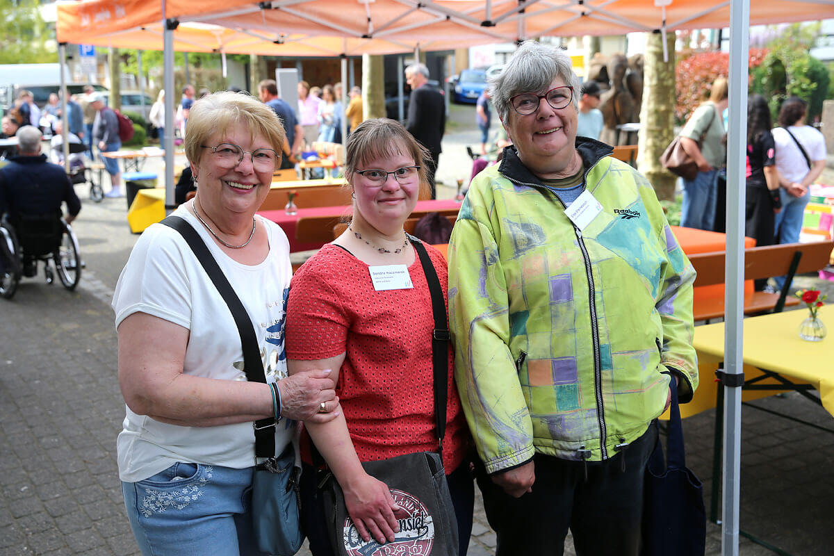 Drei Frauen stehen bei einer Veranstaltung im Freien unter orangefarbenen Zelten und lächeln in die Kamera. Im Hintergrund sind Tische mit Menschen und bunte Dekorationen zu sehen.