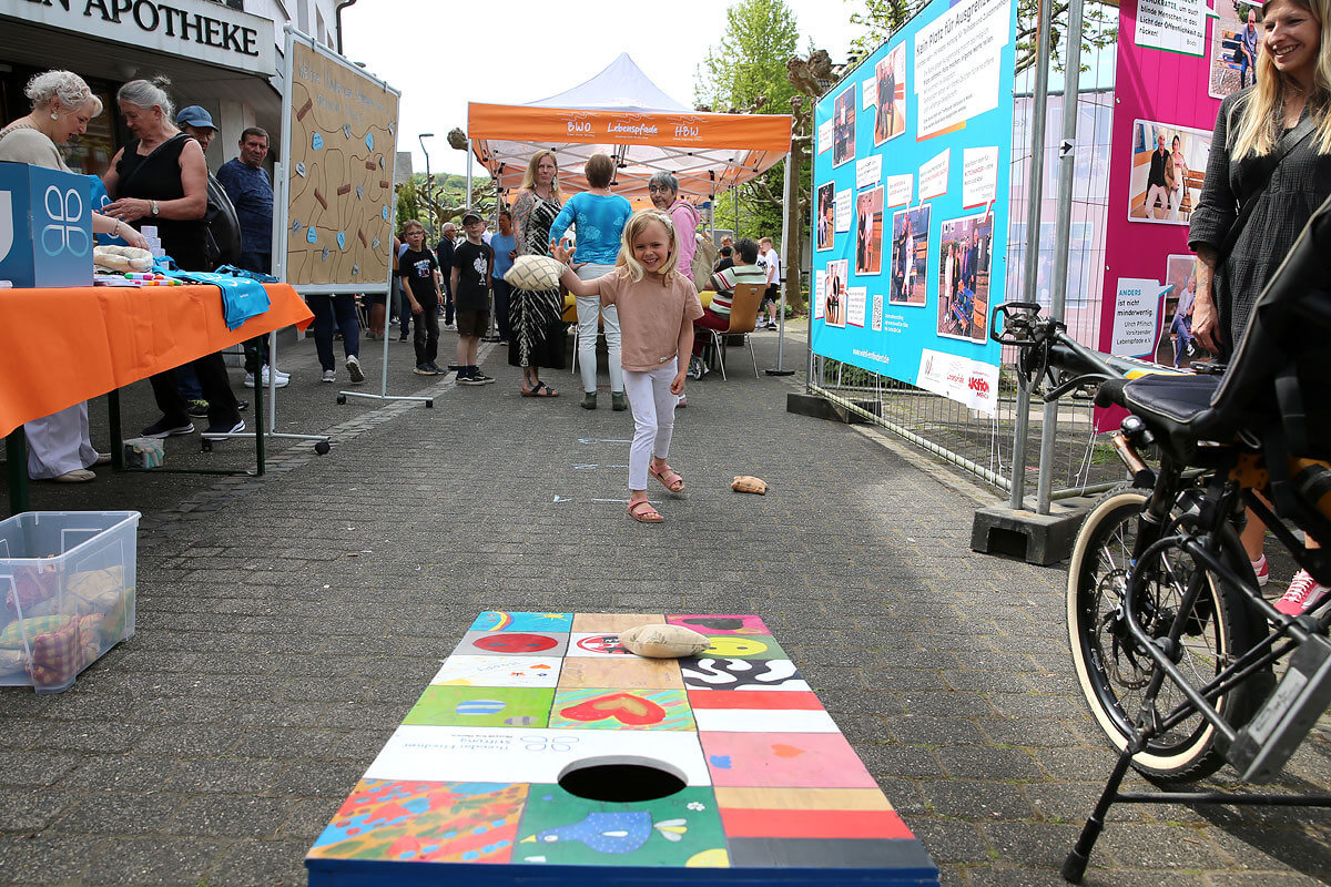 Ein junges Mädchen wirft bei einer Gemeinschaftsveranstaltung im Freien einen Bohnensack auf ein buntes Spielbrett, im Hintergrund sind Menschen und Ausstellungsstände zu sehen.