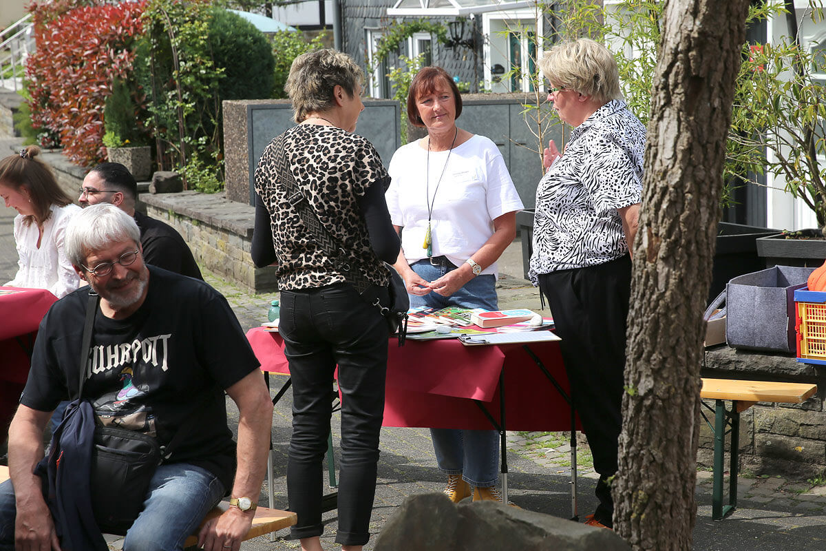 Eine Gruppe von Menschen unterhält sich an Tischen im Freien; drei Frauen stehen neben einem Ausstellungsständer mit Zeitungen, während andere in der Nähe sitzen.