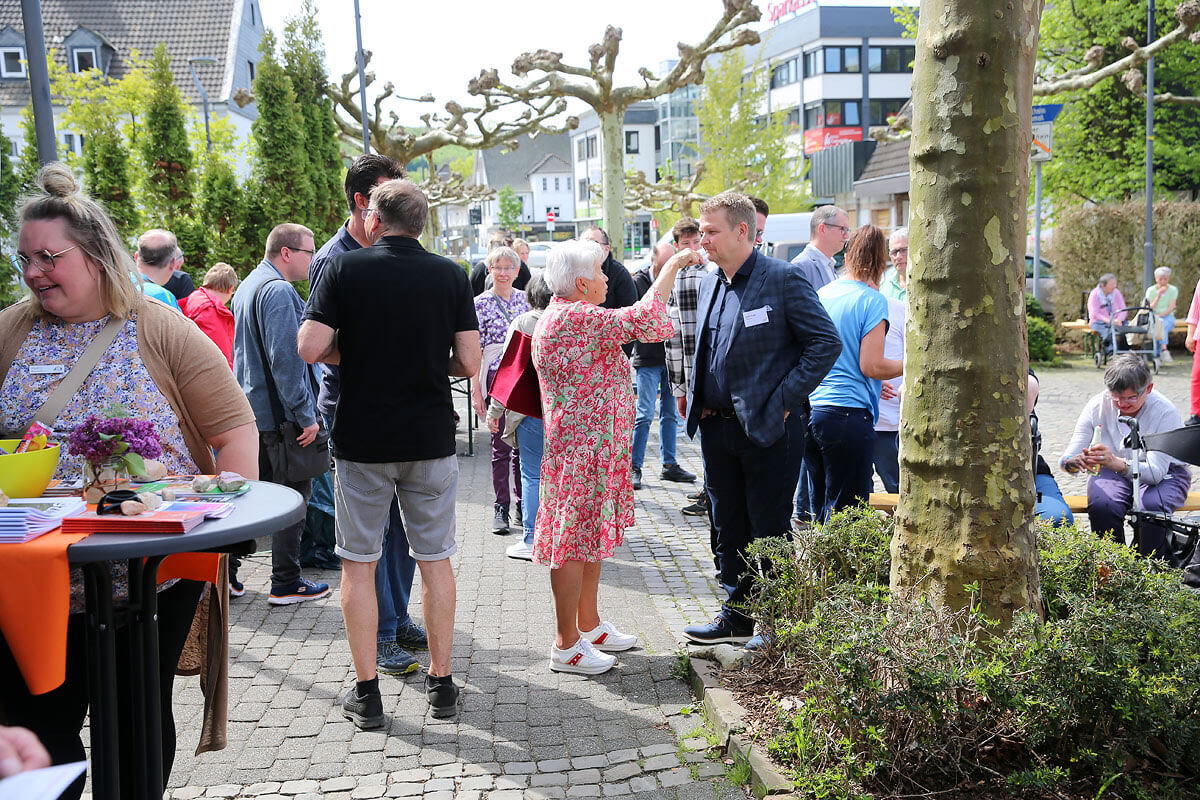 An einem sonnigen Tag versammelt sich eine Gruppe von Menschen – einige stehen, andere sitzen – im Freien auf einer gepflasterten Fläche, im Hintergrund sind Bäume und Gebäude zu sehen.