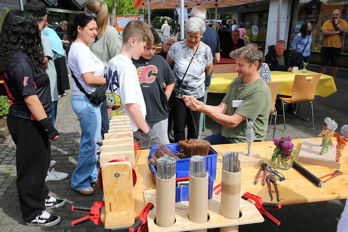 Ein Mann führt bei einer Veranstaltung im Freien einer Gruppe von Menschen vor, wie man Drahtskulpturen anfertigt. Auf einem Holztisch liegen Werkzeuge, Draht und fertige Skulpturen ausgestellt.