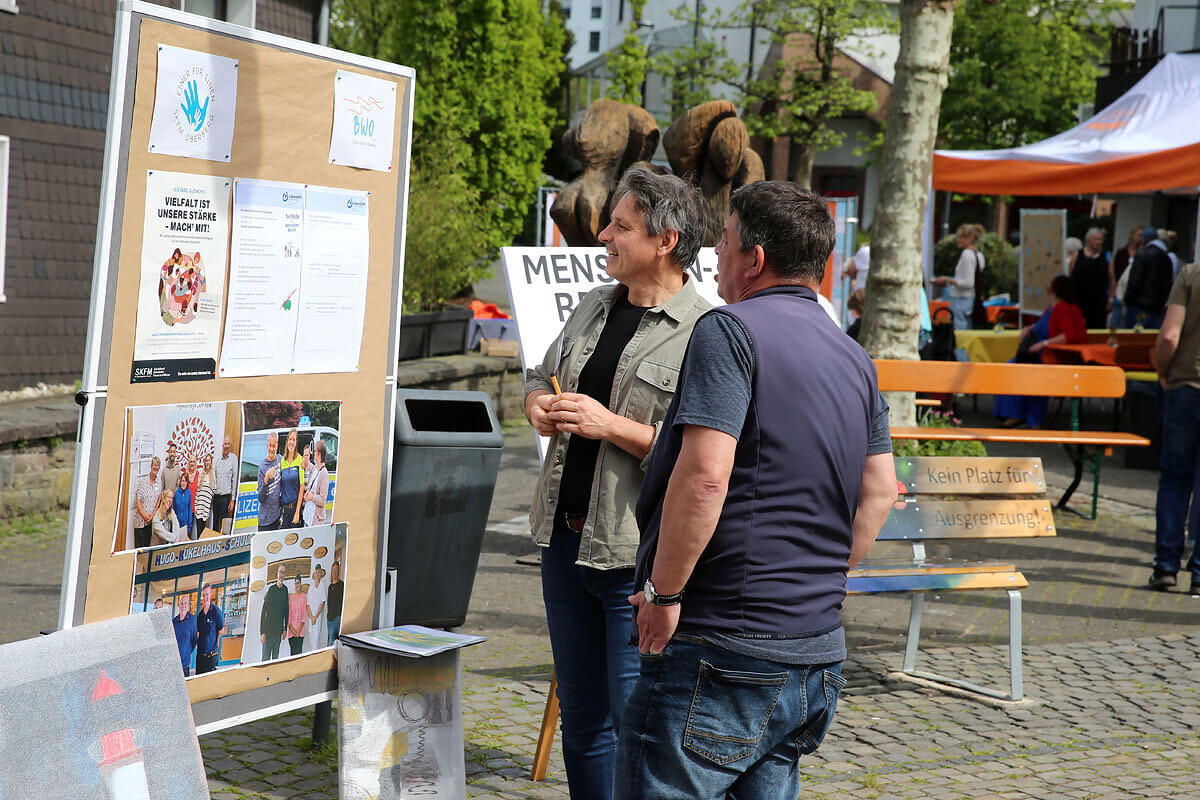 Zwei Männer stehen im Freien und schauen sich bei einer öffentlichen Veranstaltung eine Informationstafel an; im Hintergrund sind weitere Personen und Zelte zu sehen.