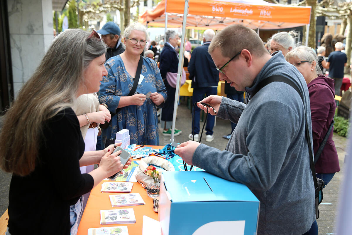 An einem Stand im Freien, an dem Broschüren ausliegen und eine Spendenbox aufgestellt ist, kommen die Menschen miteinander ins Gespräch, während andere an den nahegelegenen Ständen unter orangefarbenen Markisen stöbern.