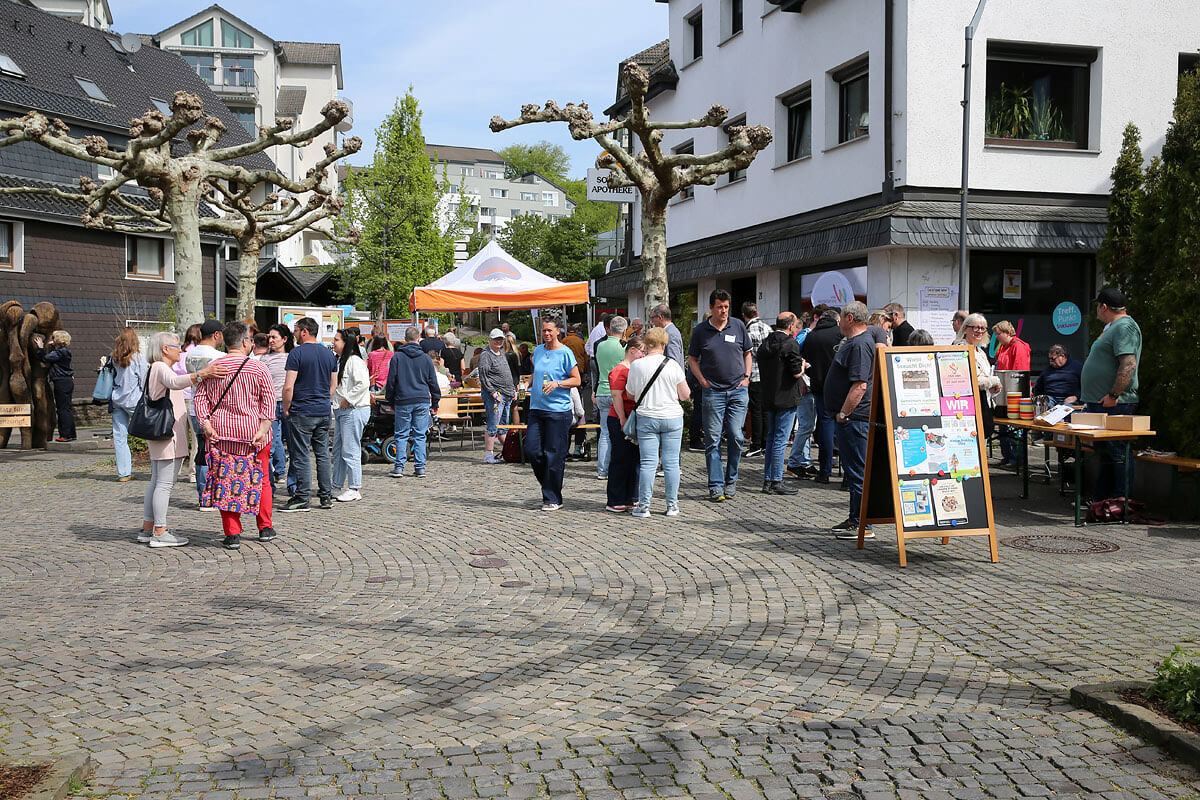 An einem sonnigen Tag versammelt sich eine Gruppe von Menschen in einem mit Kopfsteinpflaster gepflasterten Innenhof, in dem Tische, Schilder und ein orange-weißes Festzelt stehen.