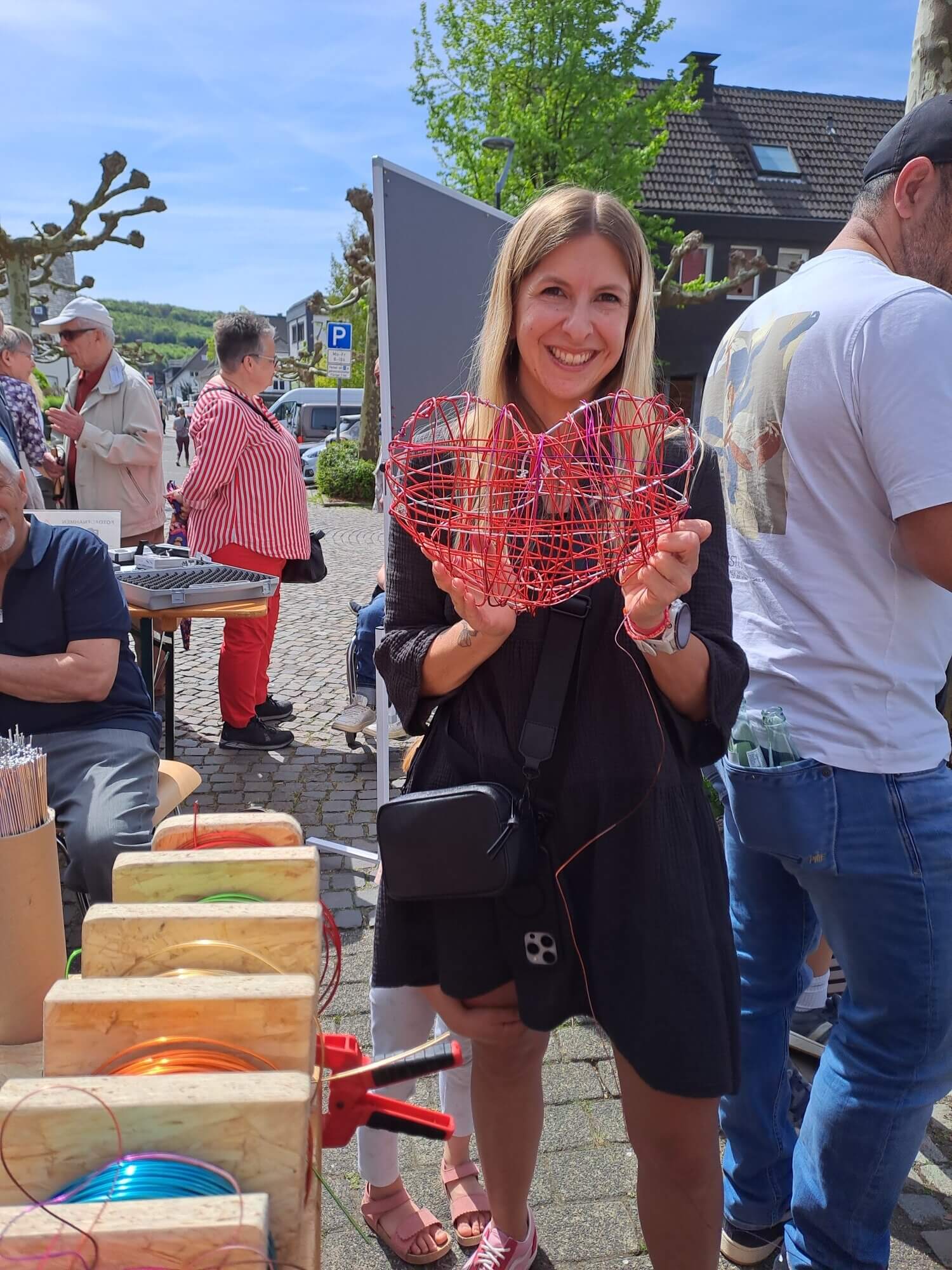 Eine Frau steht im Freien, hält eine rote Herzskulptur aus Draht in der Hand und lächelt in die Kamera. Im Hintergrund sind Menschen und Marktstände zu sehen.