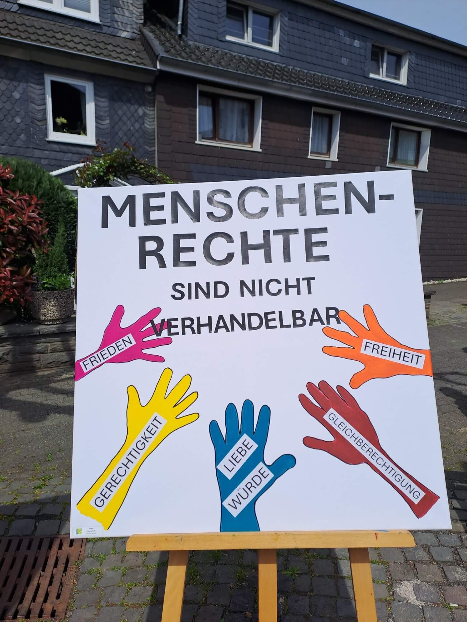 Auf einem Schild steht „Menschenrechte sind nicht verhandelbar“; daneben sind bunte Hände zu sehen, die mit den Begriffen Frieden, Freiheit, Gerechtigkeit, Gleichberechtigung, Liebe und Würde beschriftet sind. Das Schild steht im Freien in der Nähe von Häusern.