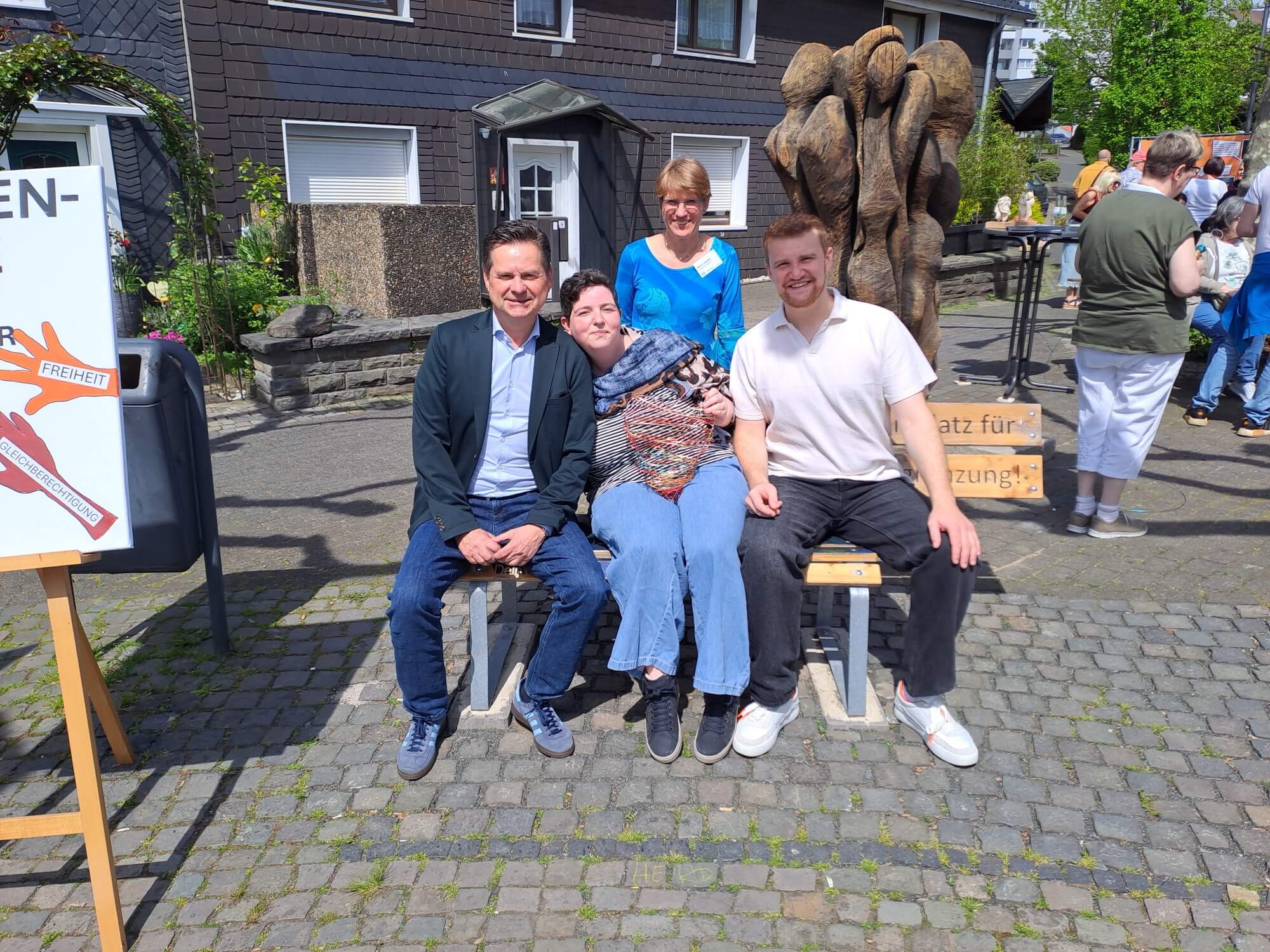 Vier Personen posieren sitzend und stehend auf einer Bank auf einem Kopfsteinpflasterplatz; im Hintergrund sind eine Holzstatue und weitere Personen zu sehen.