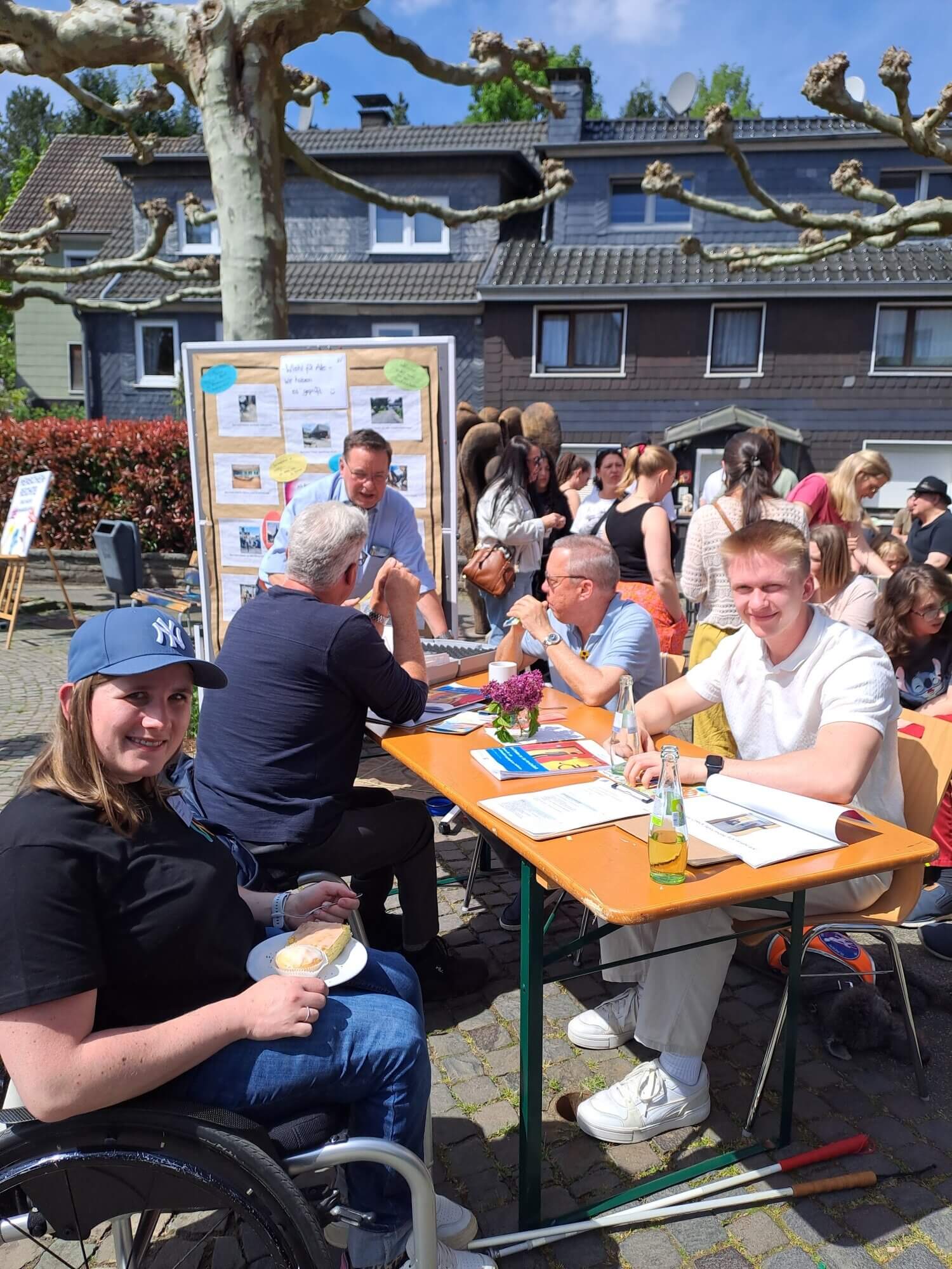Die Menschen sitzen draußen an Tischen, unterhalten sich und erledigen Papierkram, im Hintergrund sind Informationstafeln und eine Menschenmenge zu sehen. Eine Frau im Rollstuhl lächelt in die Kamera.