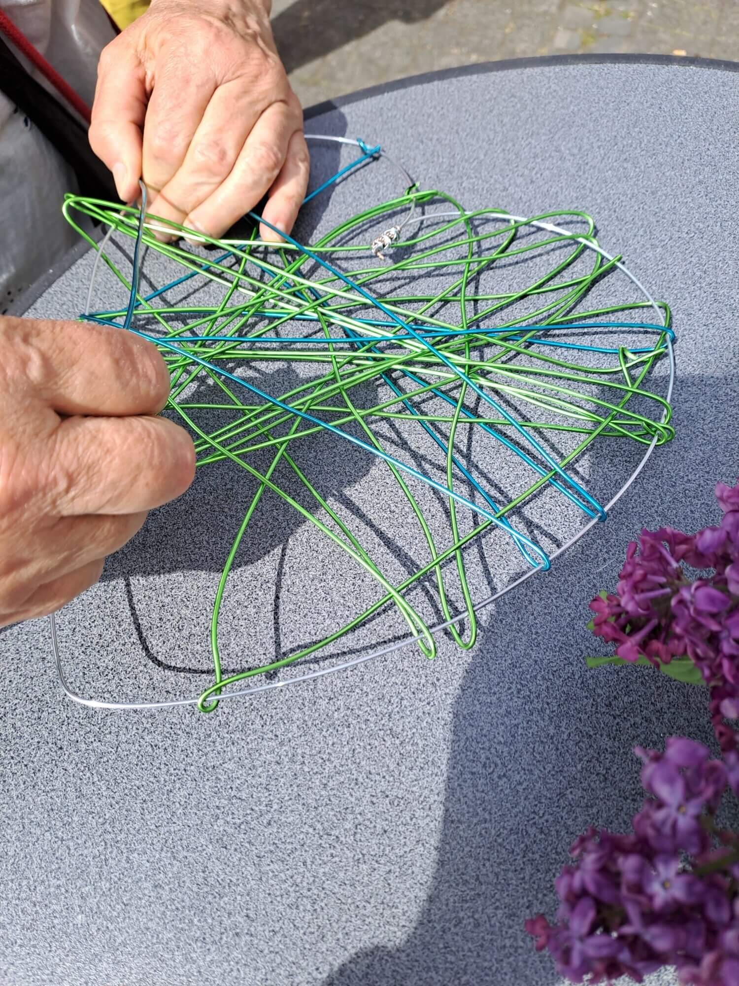 Auf einem runden, grauen Tisch im Freien biegen und formen zwei Hände bunte Drähte. In der rechten unteren Ecke des Bildes sind violette Blumen zu sehen.