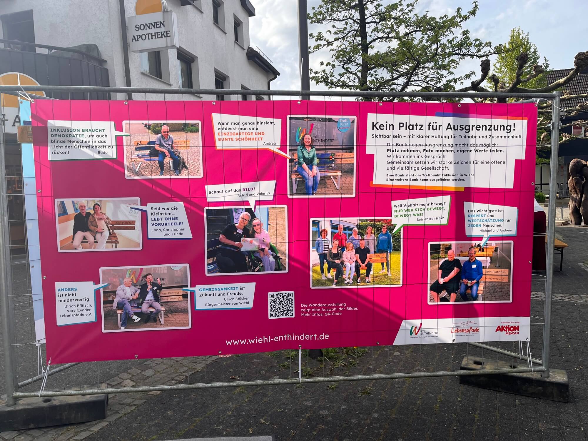 An einem Metallzaun an einer Straße in der Stadt ist ein großes rosa Transparent angebracht, auf dem Fotos von Menschen mit Behinderungen sowie Texte zu sehen sind, die sich für Inklusion einsetzen.