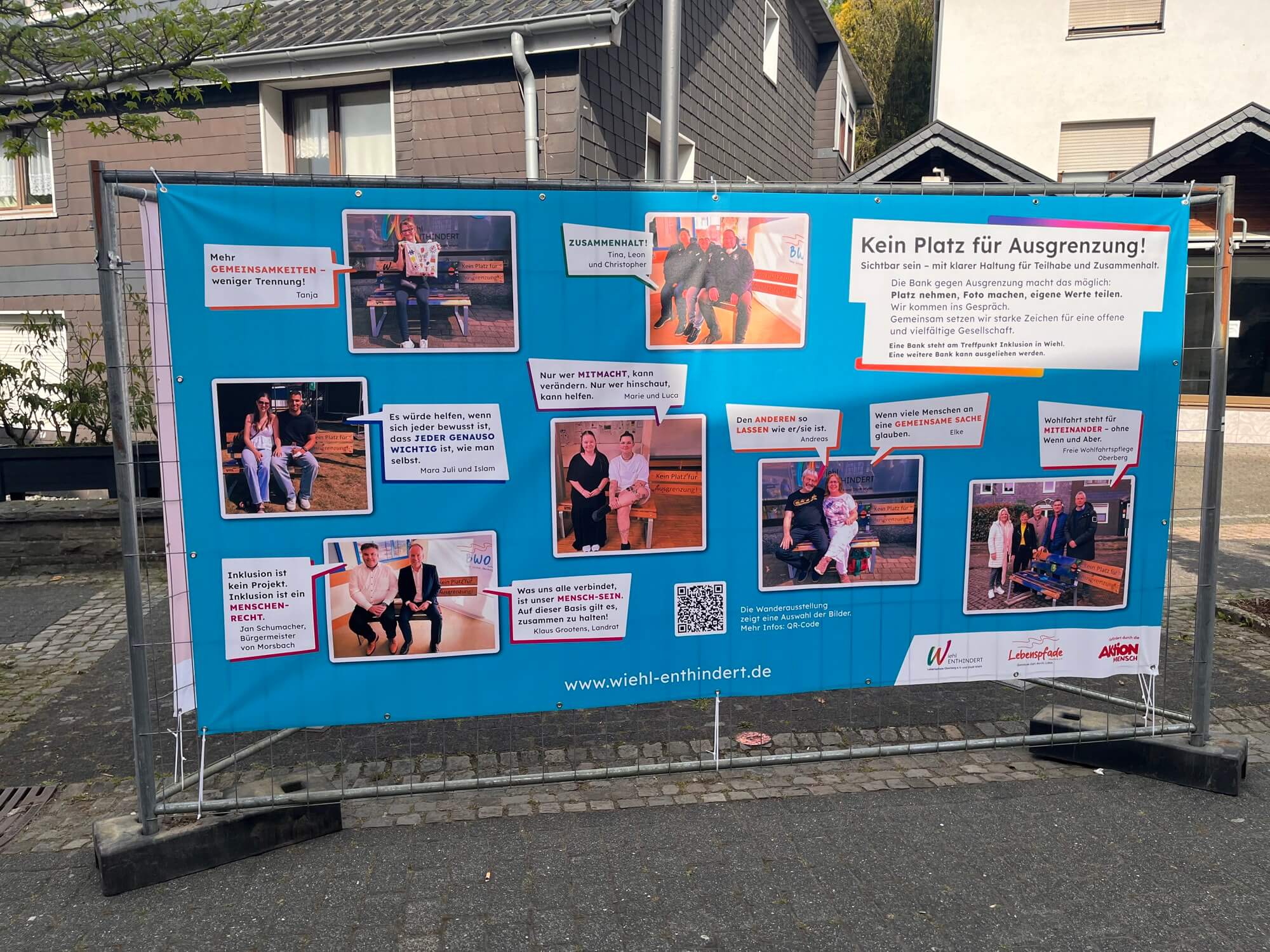 Ein Banner mit Fotos und deutschem Text zum Thema Inklusion, auf dem Menschen in verschiedenen Situationen zu sehen sind; es ist an einem Zaun entlang einer Straße angebracht, im Hintergrund sind Häuser zu sehen.