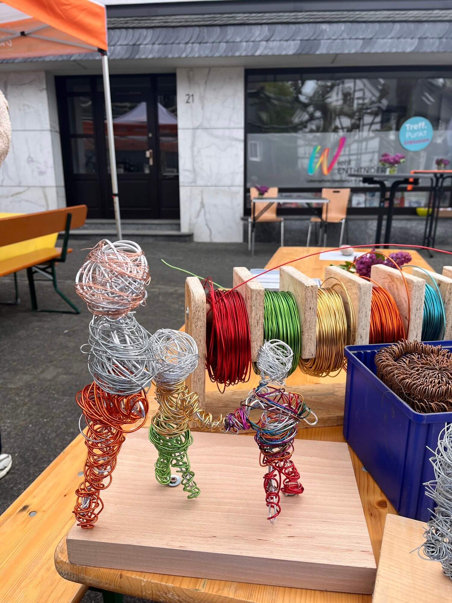 Auf einem Holzsockel sind drei aus buntem Draht geformte menschliche Figuren zu sehen, im Hintergrund sind Drahtspulen und Körbe mit Zubehör auf einem Markt oder einer Veranstaltung im Freien zu erkennen.