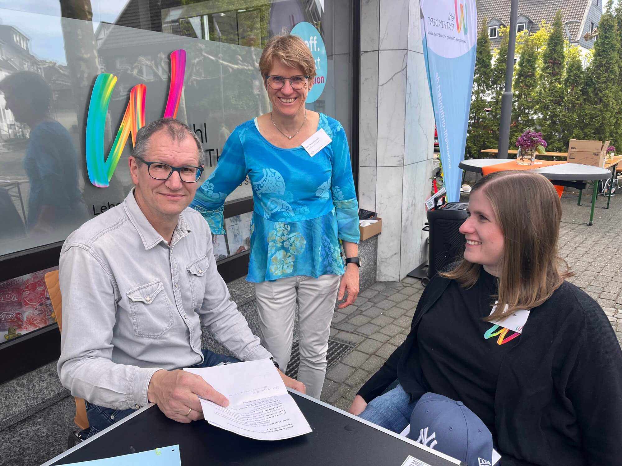 Drei Erwachsene sitzen und stehen mit Unterlagen um einen Tisch im Freien herum, vor einem Glasgebäude, hinter dem bunte Schilder und Veranstaltungsbanner zu sehen sind.