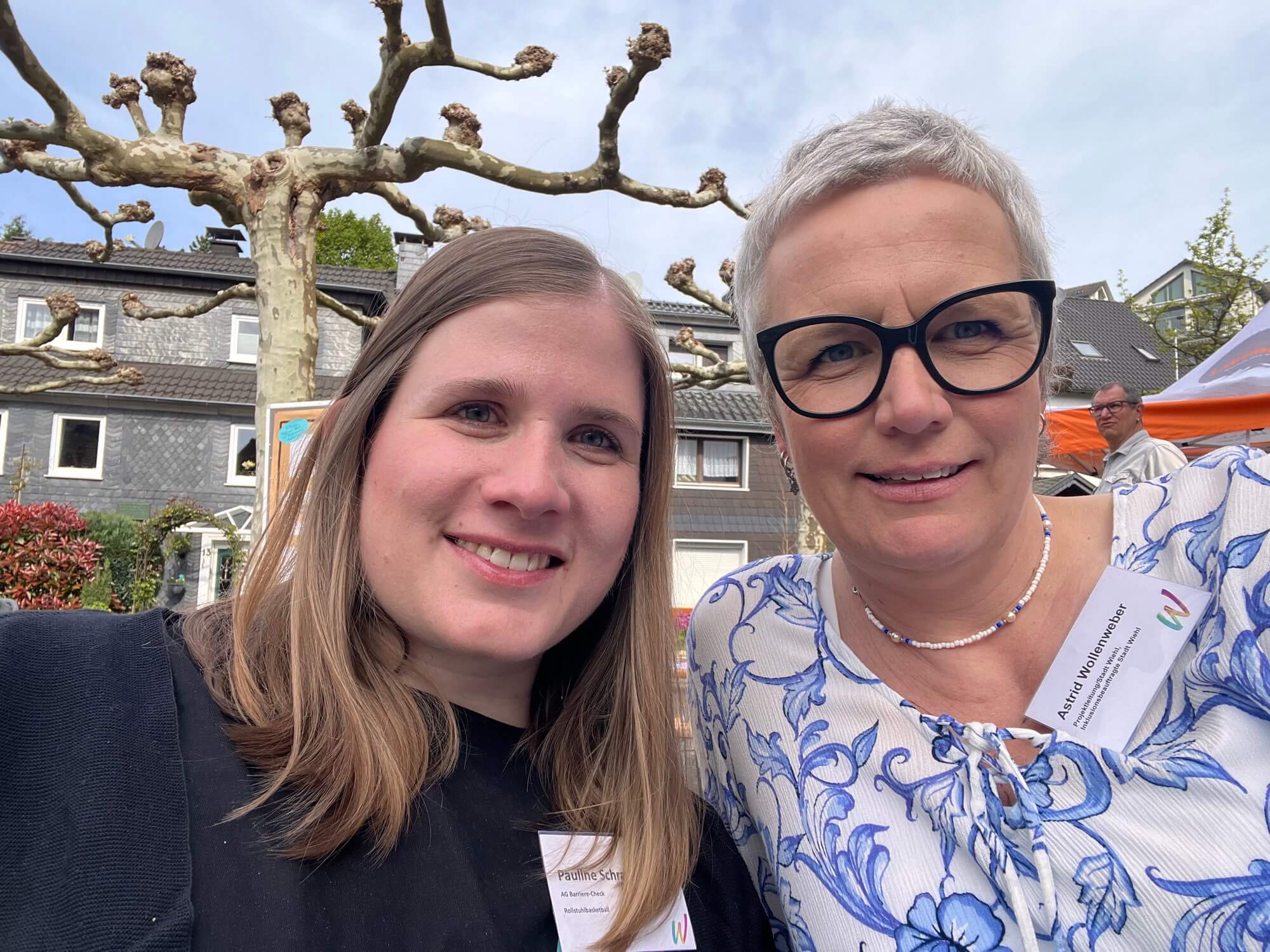 Zwei Frauen mit Namensschildern lächeln im Freien in die Kamera, im Hintergrund sind gepflegte Bäume und Häuser zu sehen.
