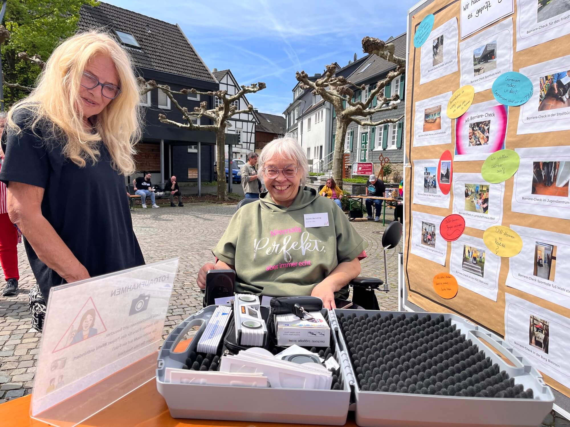 Zwei Frauen stehen an einem Stand im Freien, an dem Informationsmaterial und Ausrüstung ausgestellt sind; im Hintergrund ist eine Präsentationswand mit Fotos und Flyern zu sehen.