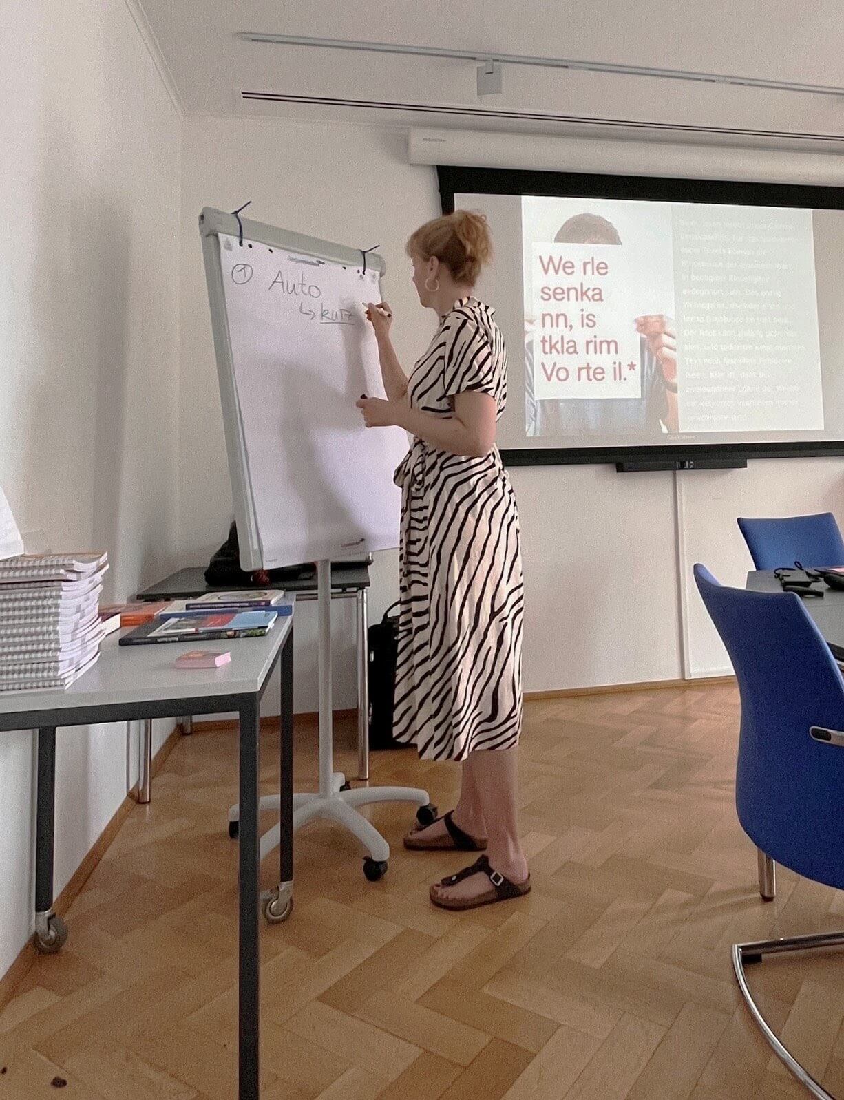 Eine Frau in einem gestreiften Kleid schreibt in einem Konferenzraum auf ein Flipchart. Im Hintergrund ist eine Projektionswand mit unscharfem Text zu sehen.