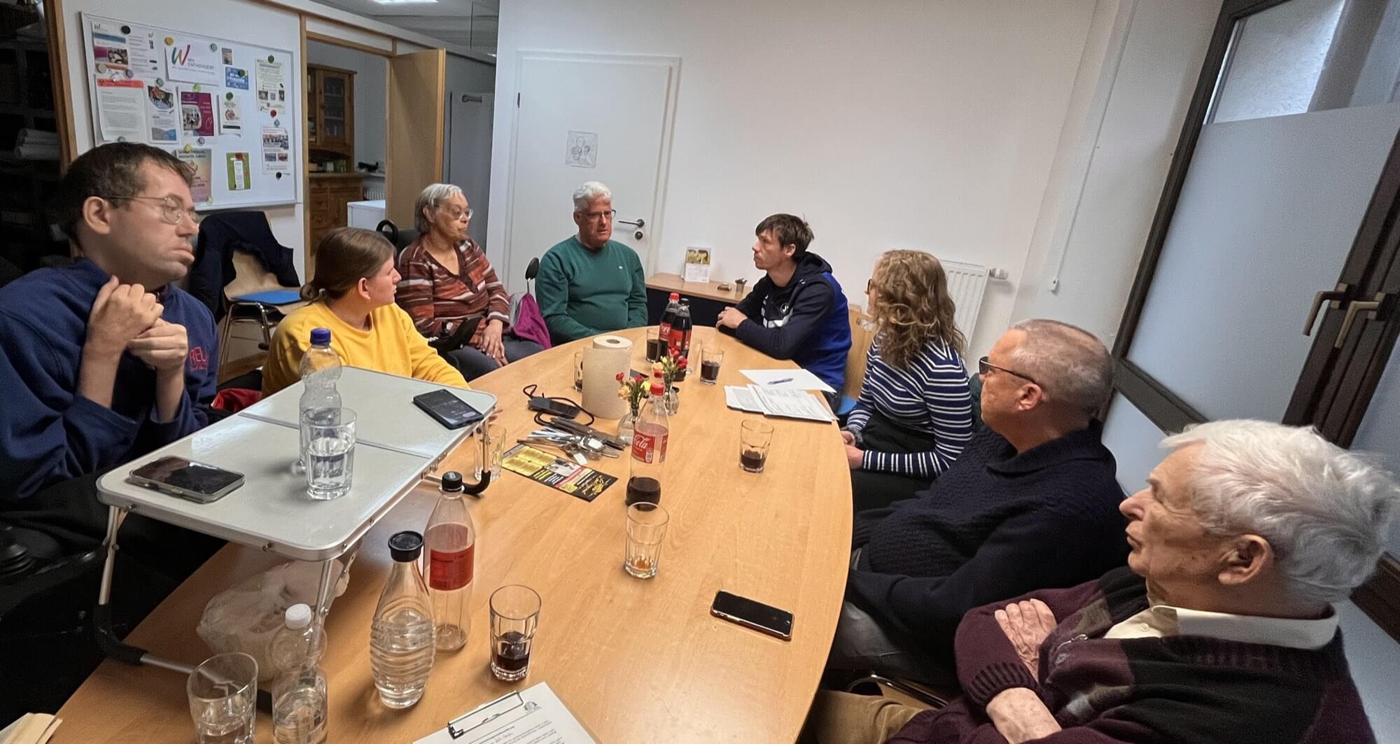 Neun Erwachsene sitzen in einem Besprechungsraum um einen Konferenztisch herum und diskutieren. Auf dem Tisch liegen verschiedene Getränke, Papiere und Stifte.
