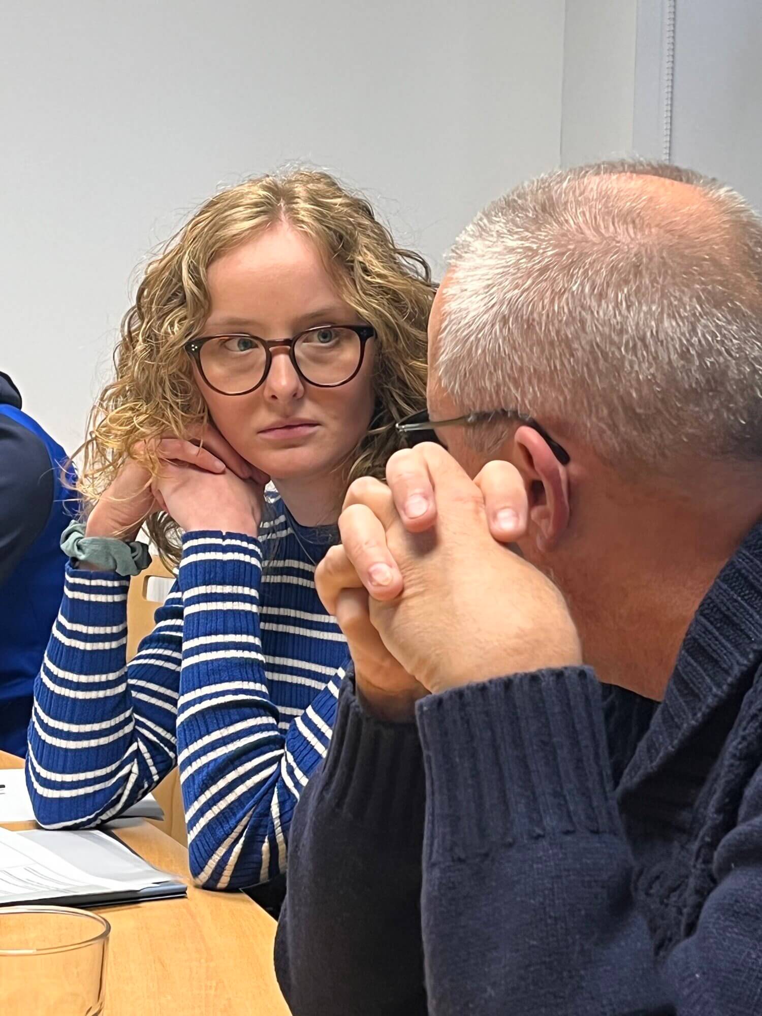 Eine Frau mit lockigem Haar und Brille hört aufmerksam einem Mann zu, der an einem Tisch in einem Besprechungsraum spricht. Auf dem Tisch liegen Papiere und ein Glas.