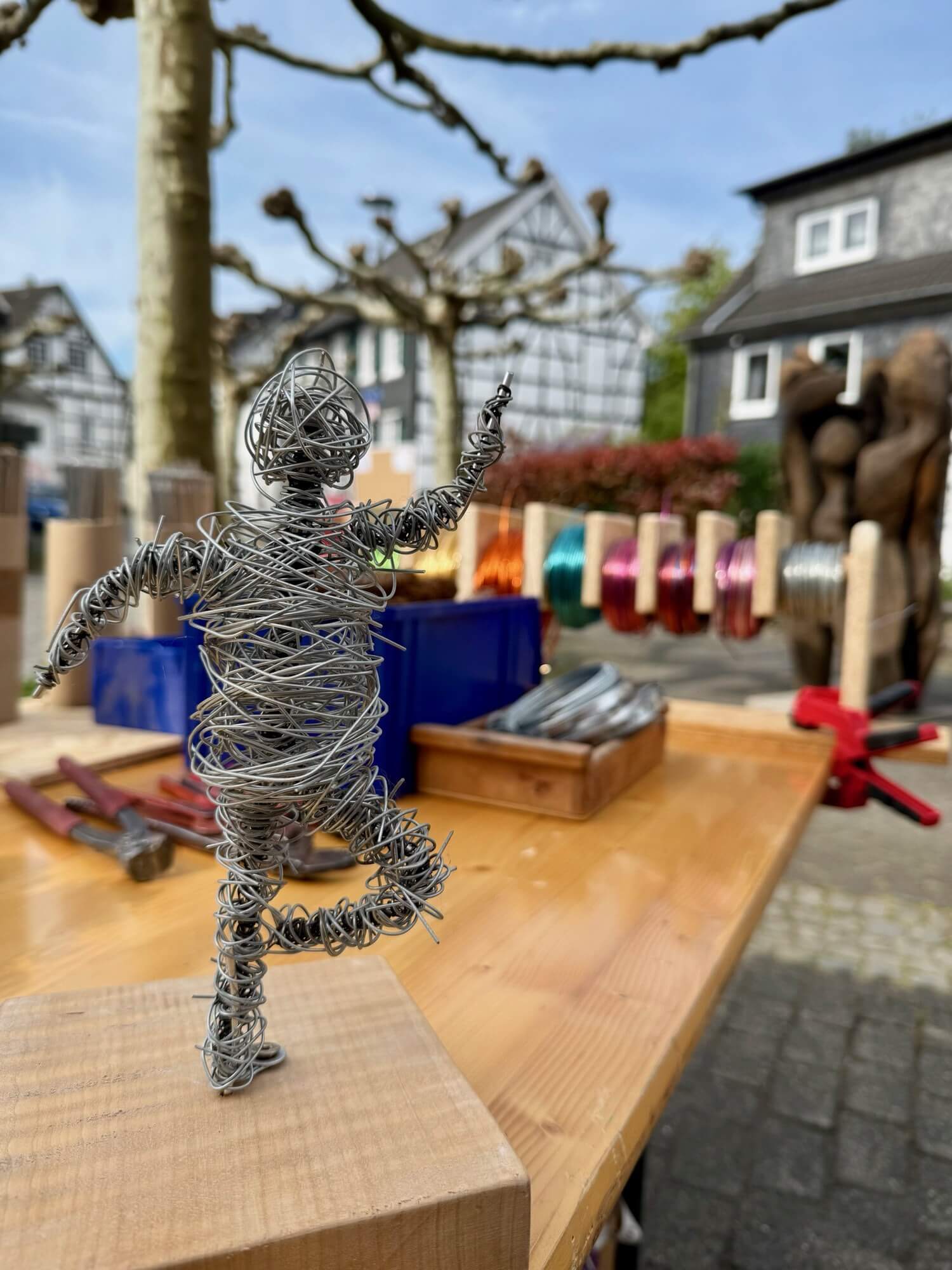 Auf einem Holztisch im Freien steht eine Drahtskulptur einer rennenden Figur, im Hintergrund sind Drahtspulen, Werkzeuge und Fachwerkhäuser zu sehen.