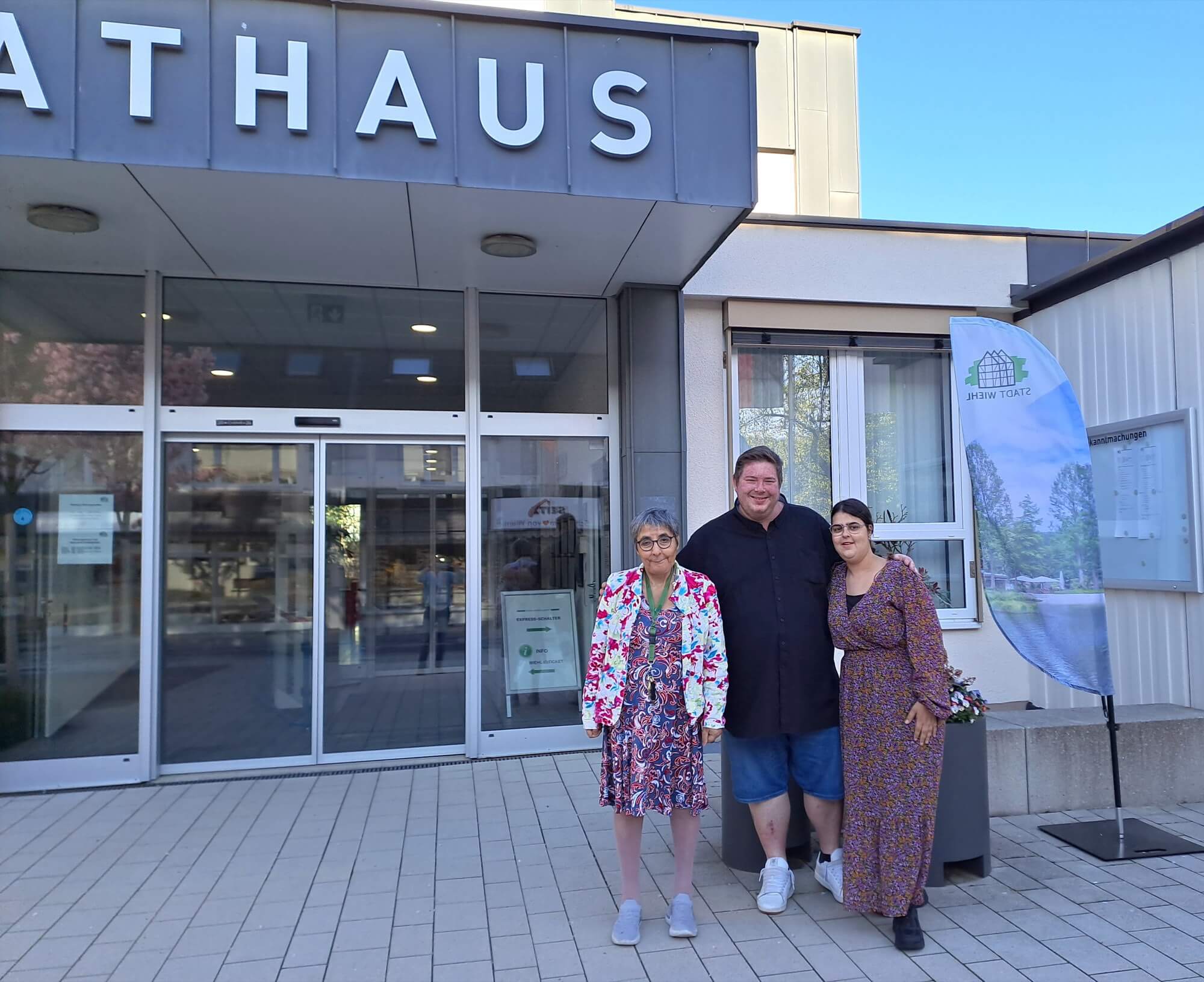 Drei Personen stehen vor dem Eingang eines modernen Gebäudes, über dessen Türen das Wort „RATHAUS“ zu lesen ist und auf dessen rechter Seite ein Werbebanner hängt.