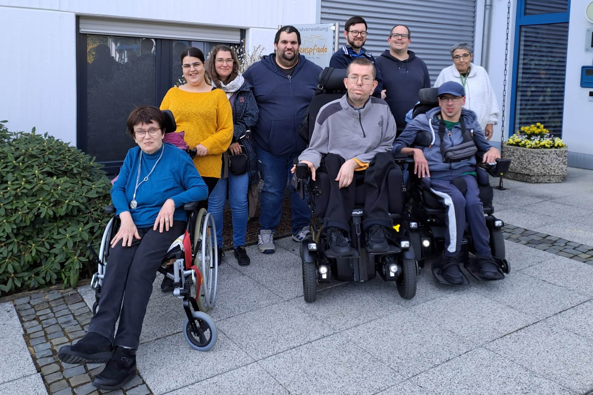 Eine Gruppe von zehn Erwachsenen, von denen einige im Rollstuhl sitzen und andere stehen, posiert gemeinsam draußen auf einer gepflasterten Fläche vor einem Gebäude.