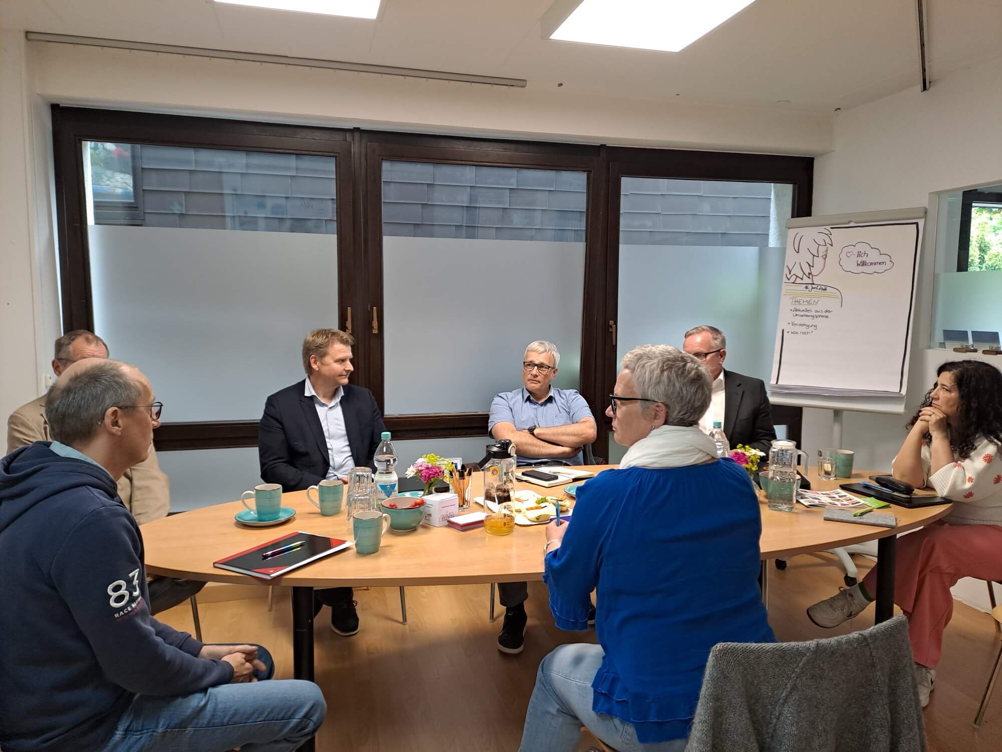 Sieben Personen sitzen in einem Büro um einen Tisch herum und halten eine Besprechung ab; dabei haben sie Getränke, Notizbücher und Snacks dabei; im Hintergrund steht ein Flipchart mit Notizen.