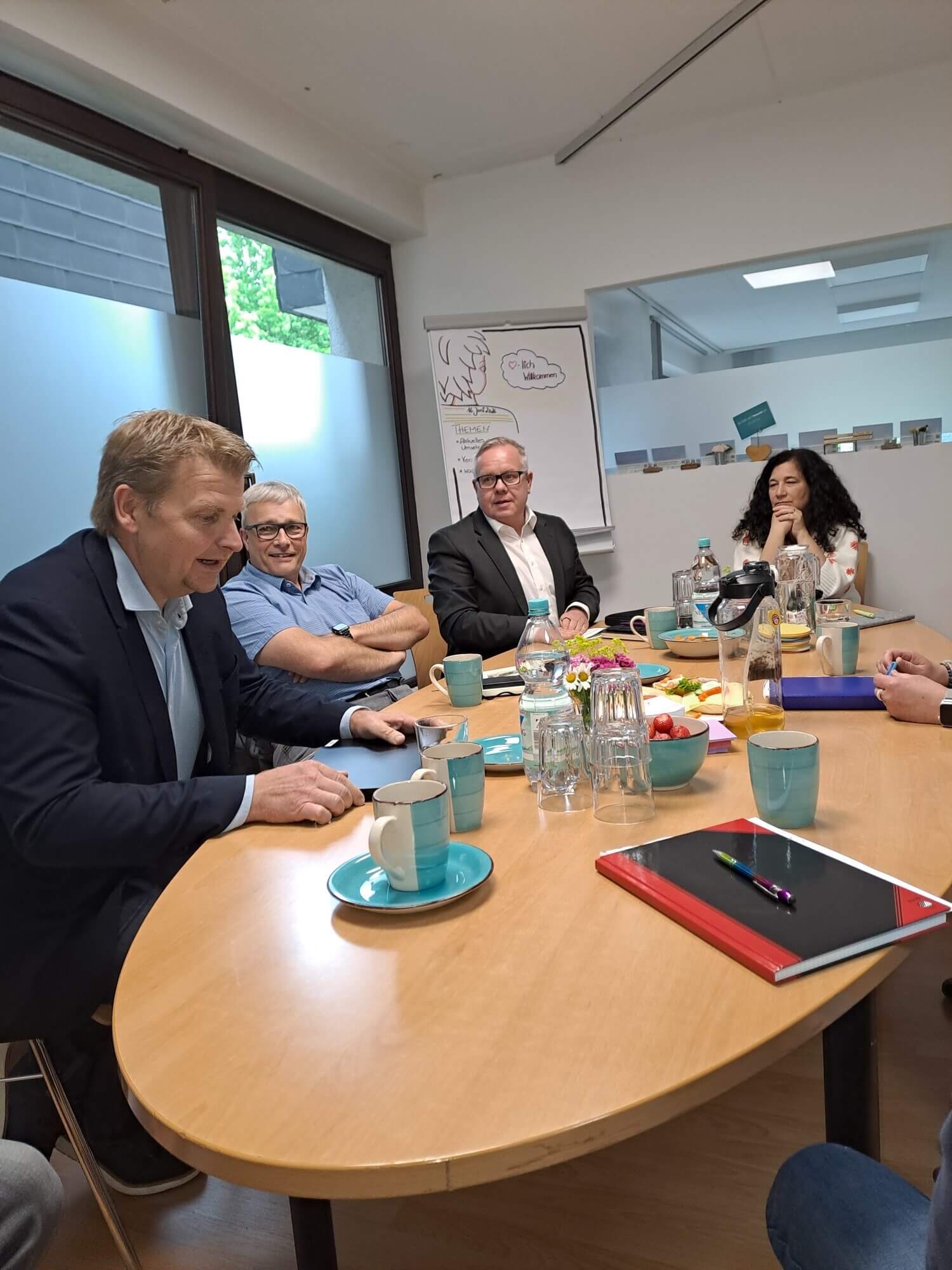 Fünf Personen sitzen mit Getränken, Snacks und Notizbüchern um einen Konferenztisch herum und diskutieren in einem modernen Besprechungsraum.