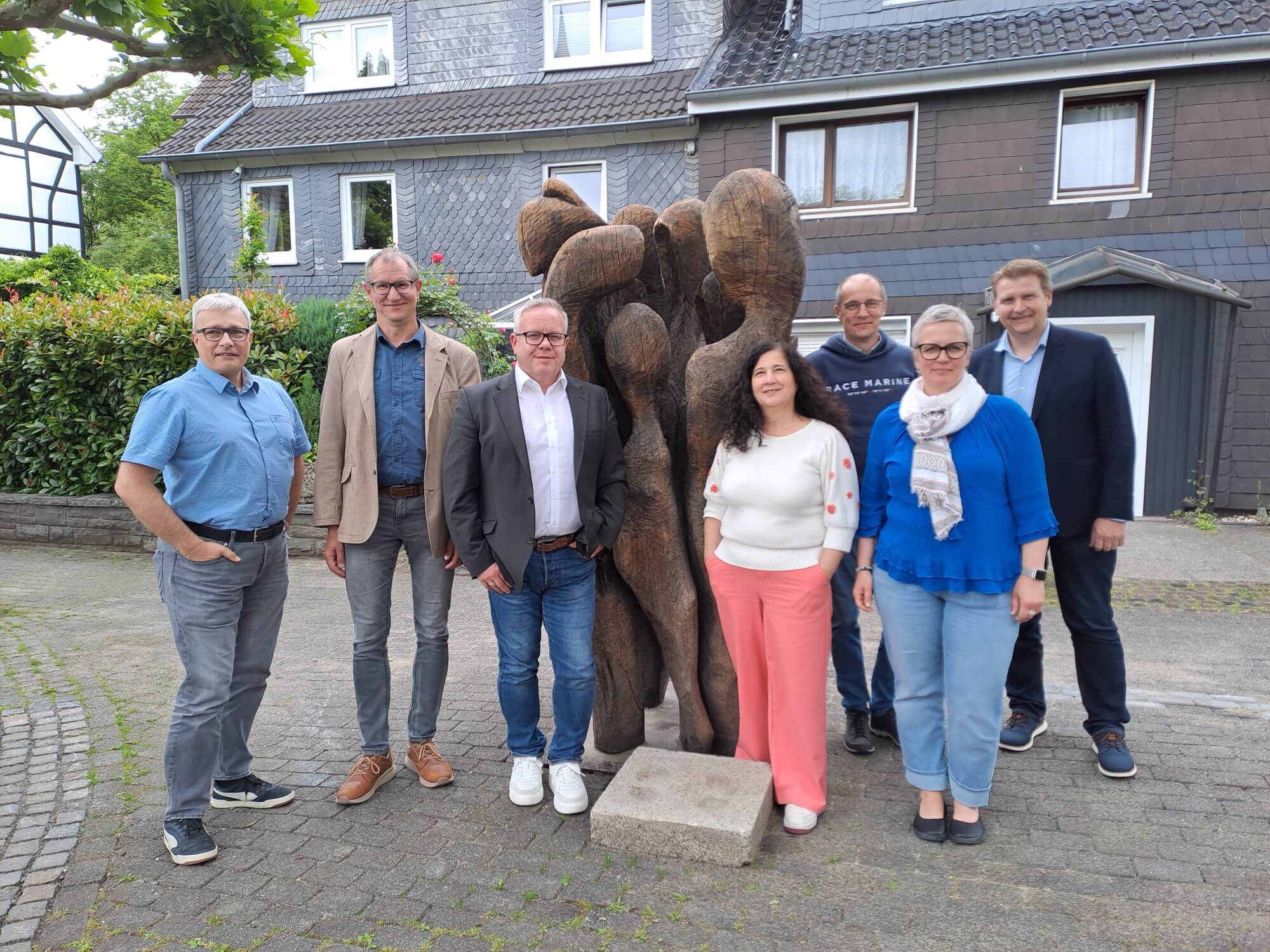 Sieben Erwachsene stehen vor einer Holzskulptur mit abstrakten menschlichen Figuren vor grauen Gebäuden mit Schindeldach und posieren für ein Gruppenfoto.