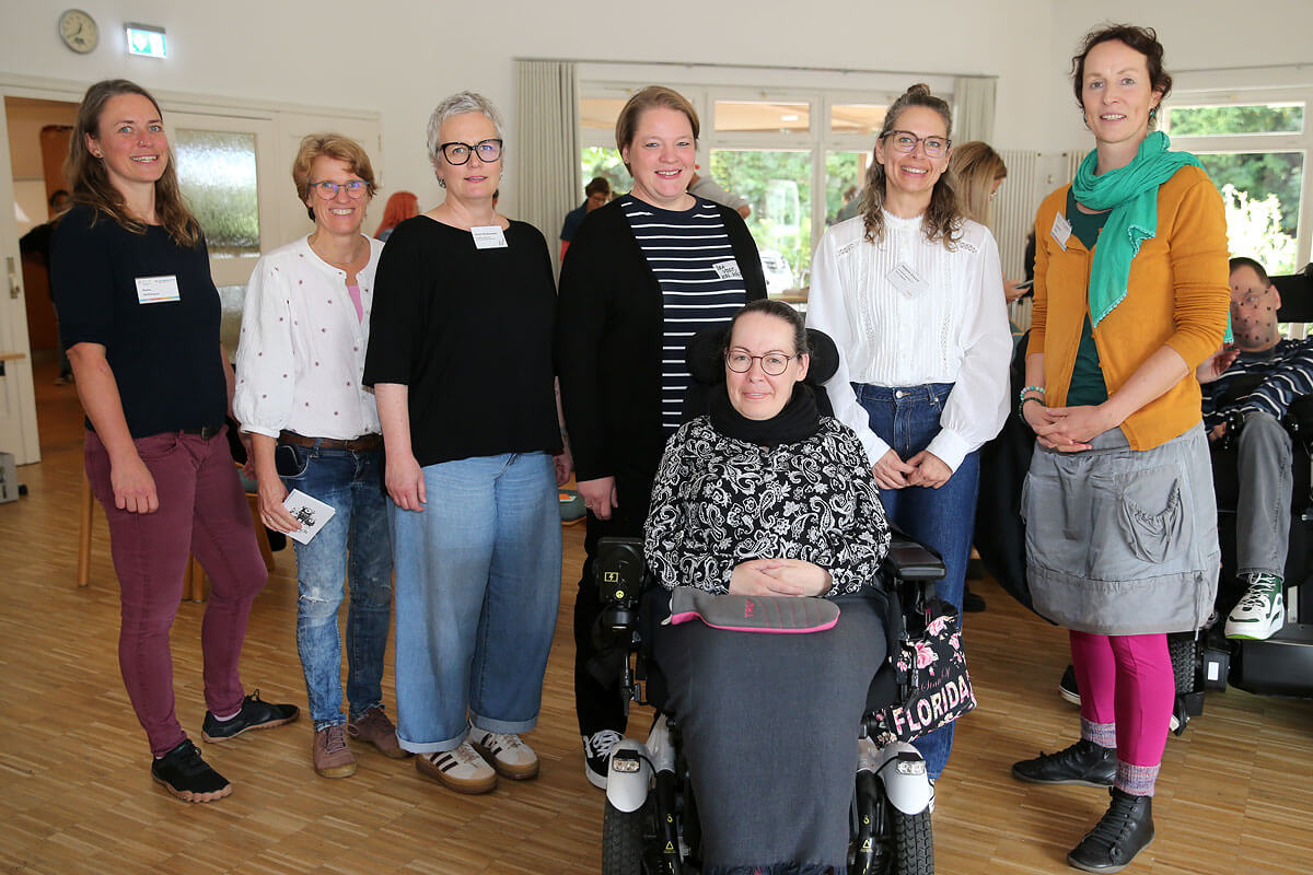 Sieben Personen posieren in einem Raum für ein Gruppenfoto, darunter eine Person im Rollstuhl. Sie lächeln und sind leger gekleidet. Der Raum hat Holzböden und weiße Wände.